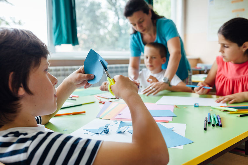 A child cutting a shape out of blue paper with scissors, coloured paper and pens spread across the table.